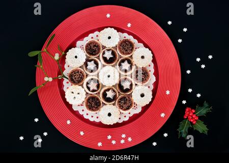 Mince torte fatte in casa per Natale su un piatto rosso con agrifoglio di bacche invernali, mistletoe e fiocchi di neve decorativi su sfondo nero. Composizione di cibo di festa Foto Stock