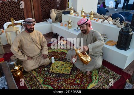 Abu Dhabi, Emirati Arabi Uniti - 2019. Due uomini arabi vestiti tradizionalmente, bevendo caffè da una scintillante caffettiera, seduti sul tappeto colorato Foto Stock