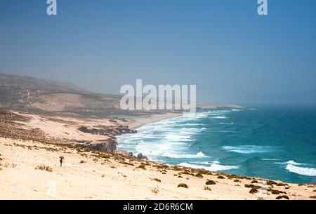 Vista dall'alto della spiaggia di Tamri. Donna cammina attraverso un campo di sabbia verso l'oceano atlantico con le onde. Foto Stock