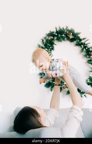 Il primo Christmas.MOM del bambino giace sul letto e tiene un ragazzino tra le braccia sullo sfondo di Una parete bianca con una corona di Natale t Foto Stock