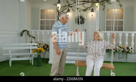 Coppia caucasica senior insieme in cortile di fronte a casa. Uomo swinging donna. Felice anziana famiglia di nonno e nonna che riposano e si godono la comunicazione Foto Stock