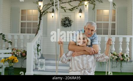 Coppia caucasica senior insieme in cortile di fronte a casa. Uomo swinging donna. Felice anziana famiglia di nonno e nonna che riposano e si godono la comunicazione Foto Stock