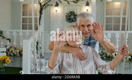Coppia caucasica senior in cortile di fronte a casa. Uomo abbracciando donna. Felice famiglia matura di nonno e nonna che sorridono, guardando la macchina fotografica, agitando le mani. Ciao saluti Foto Stock