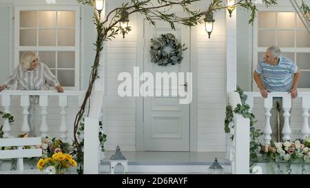 Anziano anziano coppia agitando le mani l'una all'altra mentre si trova in veranda a casa. Felice famiglia matura di nonno e nonna durante isolamento quarantena per coronavirus covid-19 Foto Stock