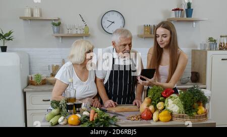 Coppia di nonni senior con nipote in cucina. Anziano uomo e donna ascolto ricetta, consigli da ragazza con tavoletta digitale di alimentazione sana. Cibo crudo mangiare dieta. Giornata mondiale vegana Foto Stock