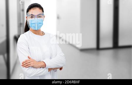 Ritratto di una dottoressa che indossa uno scudo protettivo e una maschera chirurgica, in piedi con le braccia incrociate all'ospedale corridoio. Foto Stock
