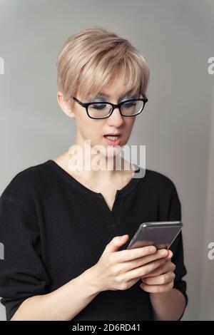 bella giovane donna bionda in occhiali guarda emotivamente allo smartphone. Foto Stock