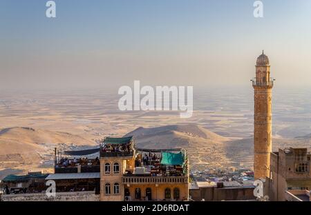 Mardin / Turchia - Ottobre 10 2020: Minareto della moschea di Mardin Ulu Cami e persone che siedono nella caffetteria sul tetto, vista della Valle della Mesopotamia Foto Stock