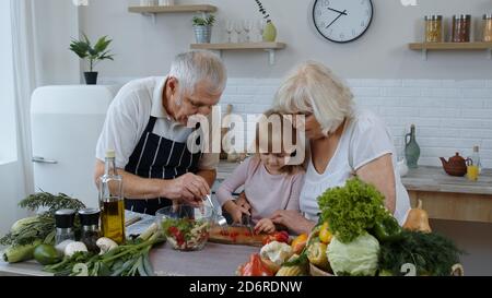 Coppia anziana in cucina insegnando alla nipote come cucinare, tagliando il pepe con il coltello. Insalata di cucina di nonna e nonno anziana con verdure fresche. Felice famiglia matura sana Foto Stock