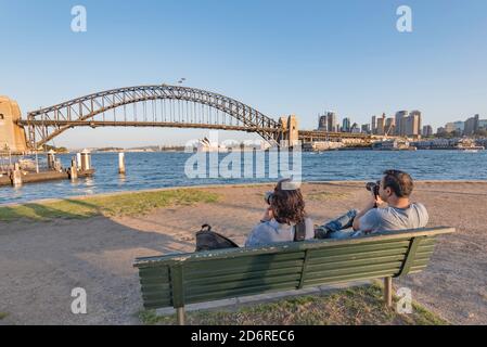 Un uomo e una donna si siedono con la macchina fotografica e scattano foto del Sydney Harbour Bridge al sole del tardo pomeriggio da McMahons Point, Sydney, Australia Foto Stock