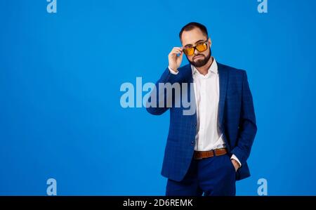 Giovane uomo che indossa occhiali da sole in un classico vestito blu sopra uno sfondo blu Foto Stock