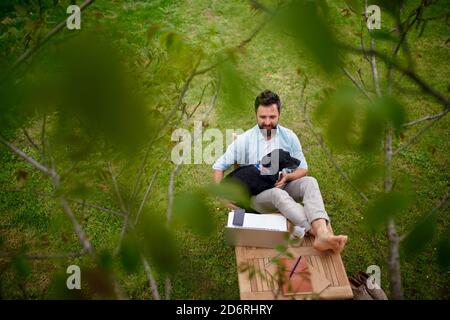 Uomo d'affari con cane e computer portatile che lavora all'aperto in giardino, concetto di home Office. Foto Stock