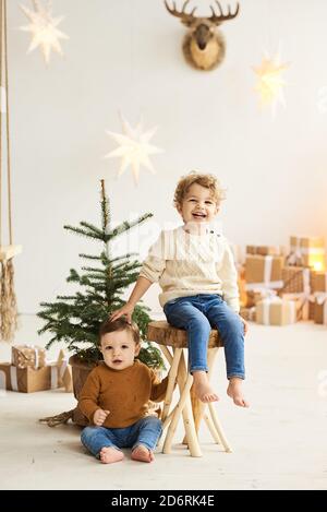 Ritratto di un piccolo bel fratello appoggiato su un legno Sedia vicino all'albero di Natale in un bianco natale decorato camera Foto Stock