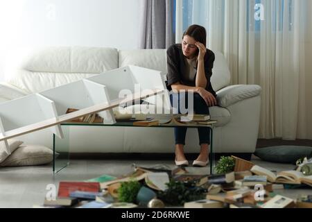 Triste inquilino che si lamenta dopo la rapina a casa seduta su un divano di notte con un soggiorno disordinato Foto Stock