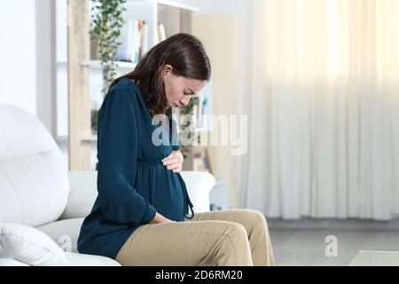 Donna incinta che soffre di mal di ventre lamentandosi di sedersi su un divano nel soggiorno di casa Foto Stock