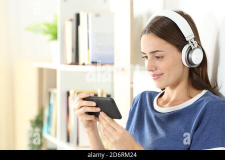 Sorridente teen che guarda i contenuti multimediali sullo smartphone indossando le cuffie in piedi nel soggiorno di casa Foto Stock