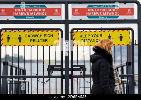 Cardiff, Galles, Regno Unito. 19 Ott 2020. Una donna che indossa una maschera facciale passa la segnaletica del coronavirus e lucchetti sulle porte del porto di Cardiff Bay come blocco nazionale di due settimane ‘Fire-break' per rallentare la diffusione del coronavirus in Galles è annunciato dal primo ministro del Galles Mark Drakeford, a partire dalle ore 1800 di venerdì 23 ottobre. Credit: Mark Hawkins/Alamy Live News Foto Stock