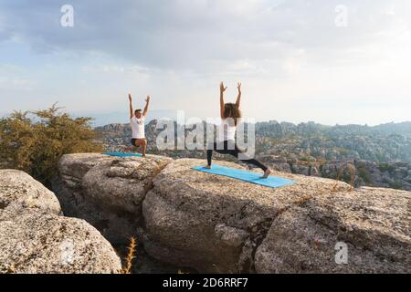 Corpo pieno di uomo e donna irriconoscibile in abbigliamento sportivo che fanno la posa Crescent Lunge mentre praticano lo yoga insieme sulla cima della roccia in montagna Foto Stock