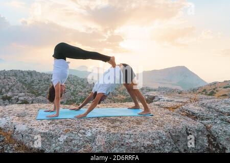 Corpo pieno di coppia irriconoscibile che pratica acro yoga e l'uomo che fa verso il basso Dog posa mentre la donna esegue handstand in cima alla roccia in moun Foto Stock