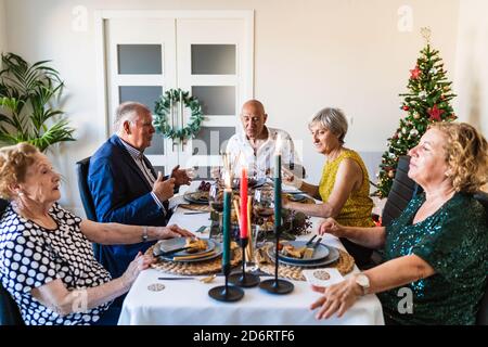 Partner che parlano a tavola con piatti gustosi e candele brucianti durante l'evento di festa vicino all'albero decorativo della pelliccia a casa Foto Stock