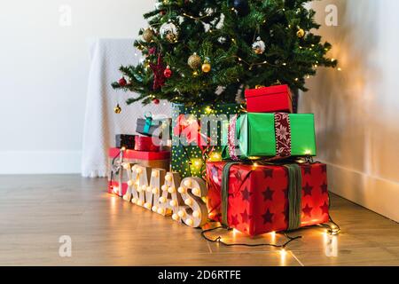 Mucchio di scatole ornamentali colorate presenti con nastri vicino decorativo pelliccia albero e lettere lucide sul pavimento con ghirlanda durante evento festivo Foto Stock