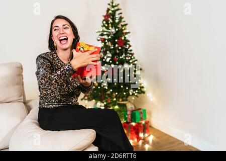 Vista laterale di una femmina eccitata in elegante scatola di apertura abiti E godendo del regalo durante la celebrazione di Natale a casa Foto Stock