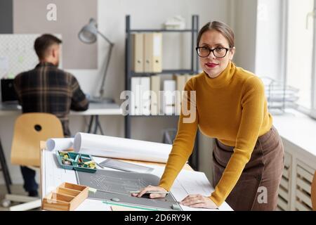Ritratto di sorridente architetto femminile disegnando progetti e piani mentre si sta in piedi alla scrivania in ufficio e guardando la fotocamera, copia spazio Foto Stock