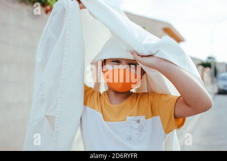 Ragazzo deliziato in costume fantasma e maschera protettiva da coronavirus Stare in città durante la vacanza di Halloween e guardare la macchina fotografica Foto Stock