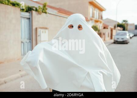 Capretto irriconoscibile in costume bianco fantasma che si diverte sulla strada Durante la vacanza di Halloween e guardando la macchina fotografica Foto Stock