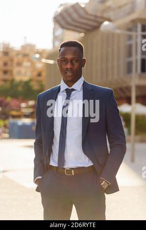 Fiducioso imprenditore africano americano maschio adulto in elegante vestito blu con laccio che guarda la fotocamera mentre si è in piedi contro città sfocate sfondo di Foto Stock