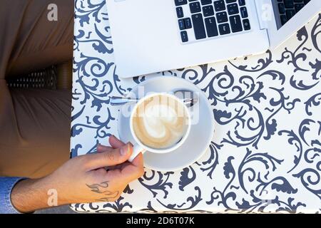 Vista dall'alto del freelancer maschile anonimo con tatuaggio seduta a mano al tavolo con una tazza di caffè e un computer portatile mentre si lavora a progetti remoti in styli Foto Stock