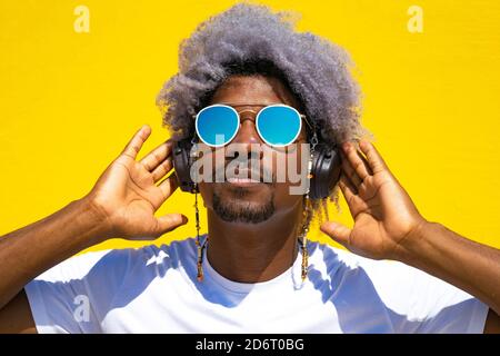 Uomo nero con capelli afro e occhiali da sole che indossano le cuffie. Concetto di ascolto di musica con le cuffie. Sfondo giallo. Foto Stock
