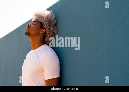 Uomo nero con capelli afro vestito di bianco appoggiato su una parete blu. Afro concetto di capelli. Uomo nero in t-shirt bianca. Foto Stock