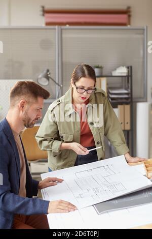 Ritratto verticale di due architetti che guardano i progetti e discutono lavorare mentre si è in piedi presso la scrivania in un ufficio moderno Foto Stock