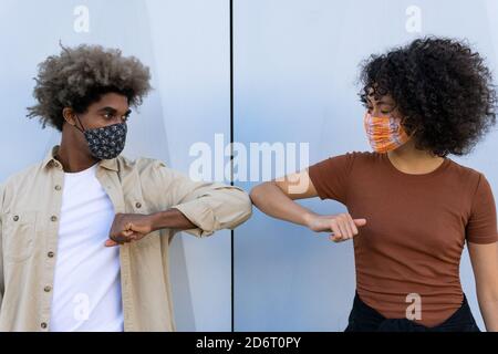 Anonimo giovane afro-americano uomo e donna con capelli ricci in abiti casual e maschere protettive che toccano i gomiti contro la parete bianca Foto Stock