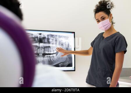 Amichevole afroamericana dentista femminile in uniforme e maschera medica Esame dei raggi X di mascelle e denti durante la condivisione delle informazioni con p non riconoscibile Foto Stock