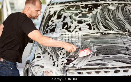 Un giovane serio che indossa una t-shirt nera sta lavando la sua auto con una spazzola e sapone all'aperto, vista laterale, primo piano Foto Stock