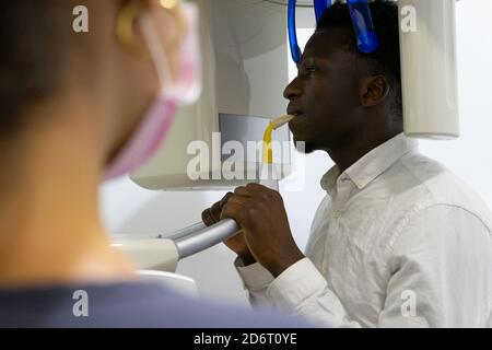 Vista posteriore del dentista irriconoscibile che prende raggi X dei denti di paziente maschio nero seduto in stanza medica in moderno clinica Foto Stock