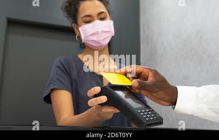 Basso angolo di African American manager femminile in maschera medica Ricezione del pagamento tramite terminale POS e carta di plastica da Crop paziente maschile in clin moderno Foto Stock