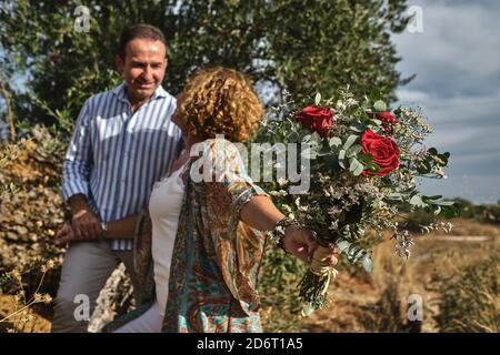 Allegra coppia di mezza età in abbigliamento casual in piedi insieme con tenero bouquet di fiori mentre vi godete una passeggiata nel giardino estivo il giorno di sole Foto Stock