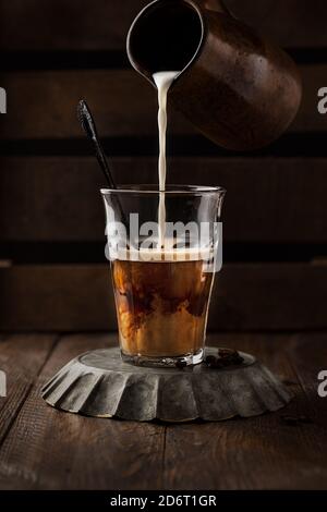 Persona anonima aggiungendo latte fresco in vetro con caffè caldo posto su un tavolo di legno in un accogliente cafe Foto Stock