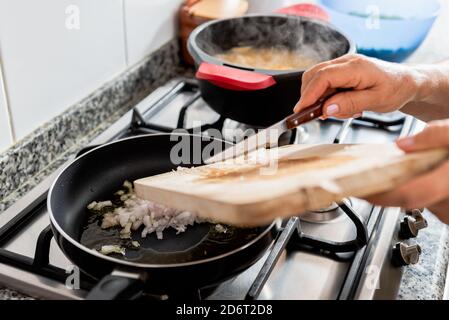 Crop anonymous Woman mettere cipolla rossa tagliata in padella calda con olio mentre preparare il tradizionale piatto di pesce catalano in cucina casa Foto Stock