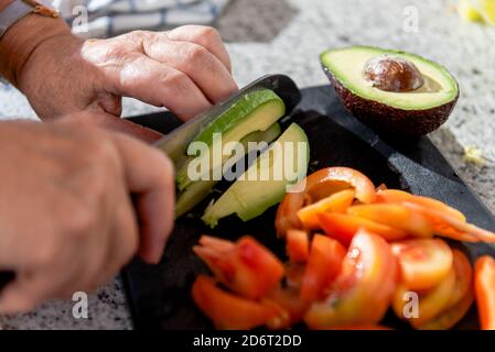 Raccolto ad angolo alto anonimo Senior femmina taglio maturo avocado organico e pomodori su tavola nera mentre cucinando in cucina soleggiata Foto Stock