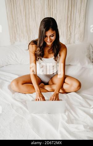 Dall'alto felicissima giovane femmina in Sleepwear seduto a letto e navigare in un computer portatile dopo il risveglio in una camera da letto leggera al mattino Foto Stock