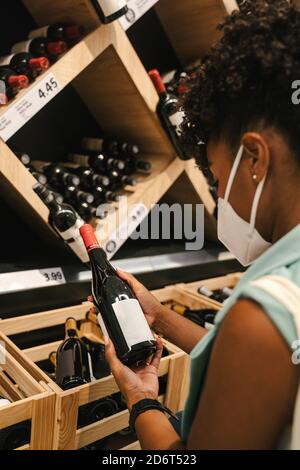 Vista laterale del cliente africano americano di prodotto in protezione maschera per la prevenzione del coronavirus che esamina una bottiglia di vino durante la scelta bevande in sup Foto Stock