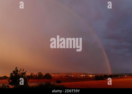 Scenario di vibrante arcobaleno che splende su un cielo scuro e drammatico coperto con spesse nuvole pesanti in serata con vasti campi agricoli in campagna Foto Stock