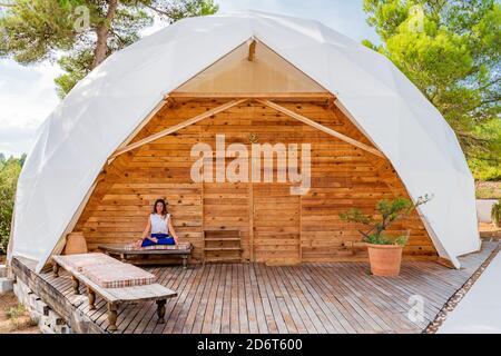 Irriconoscibile fit lady in sportswear seduto in Padmasana posa su panca vicino costruzione in legno in estate Foto Stock