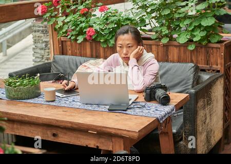 Concentrata giornalista asiatica seduta a tavola con foto digitale fotocamera e utilizzo del computer portatile durante la preparazione di un nuovo articolo Foto Stock