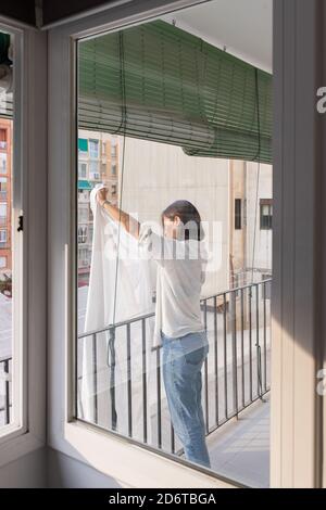 Vista laterale della giovane femmina in camicia bianca e jeans lavanderia appesa su stendibiancheria sul balcone Foto Stock