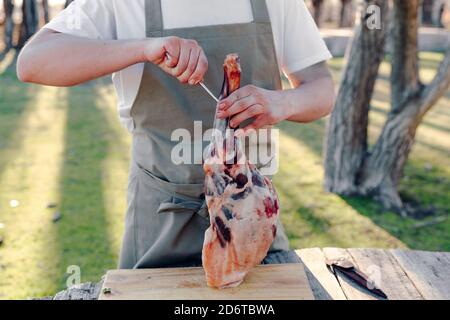 Anonimo cuoco maschile in grembiule in piedi a tavola in campagna con gamba grezza di agnello e preparazione di carne per appendere su gancio in metallo Foto Stock